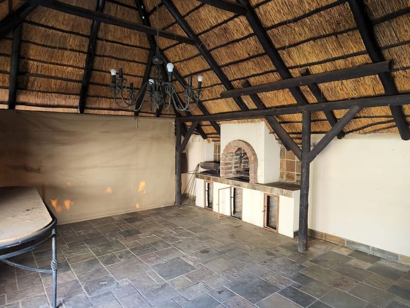 Covered outdoor entertainment area featuring a traditional thatched roof, exposed timber beams, stone flooring, and a built-in brick braai fireplace.