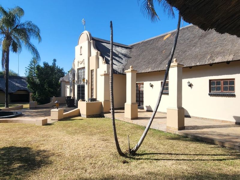 Rear view of a Cape Dutch style house featuring a traditional thatched roof, expansive paved patio area, and a central water fountain in the garden.