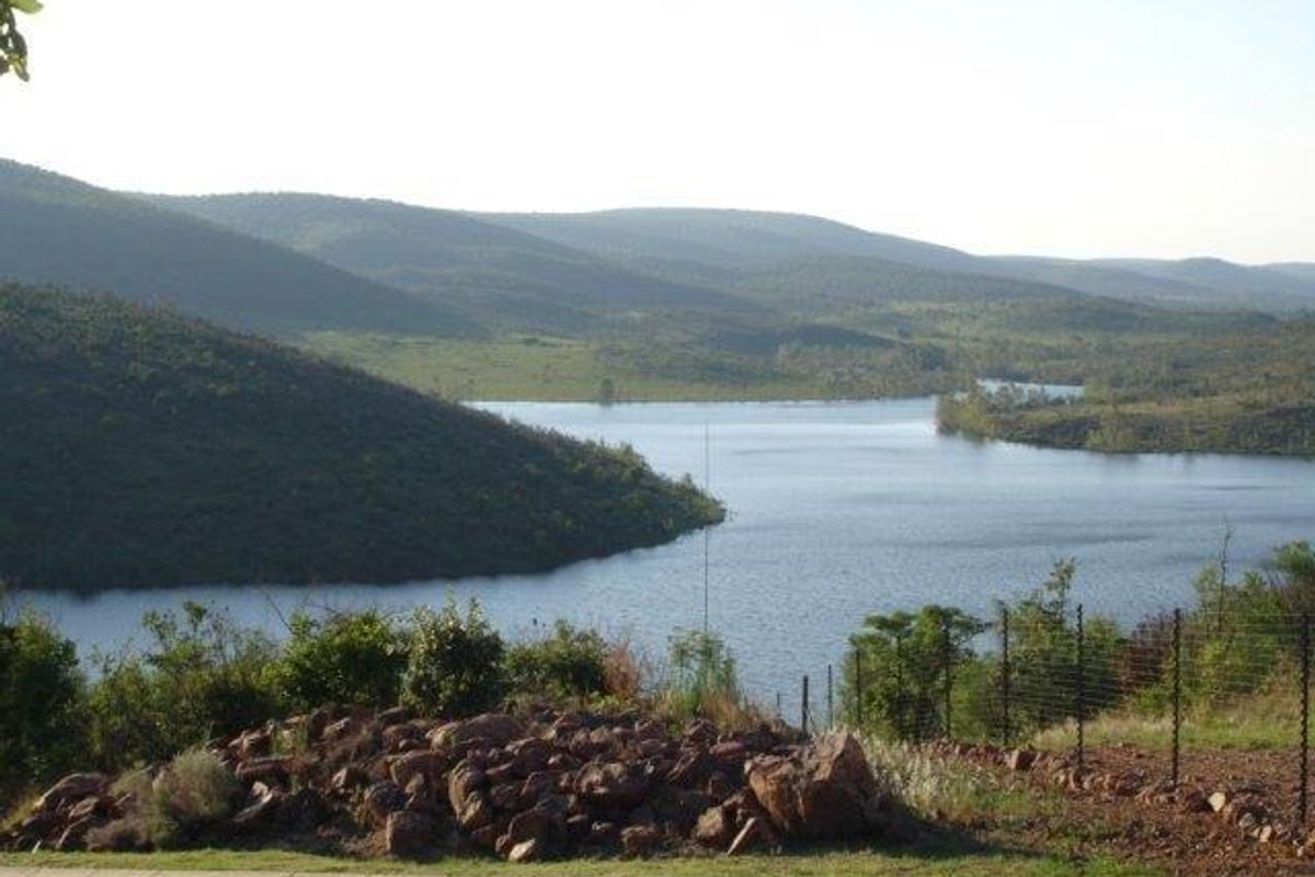 A scenic view overlooking a large dam or lake surrounded by rolling hills and natural vegetation, captured from the edge of a residential property.