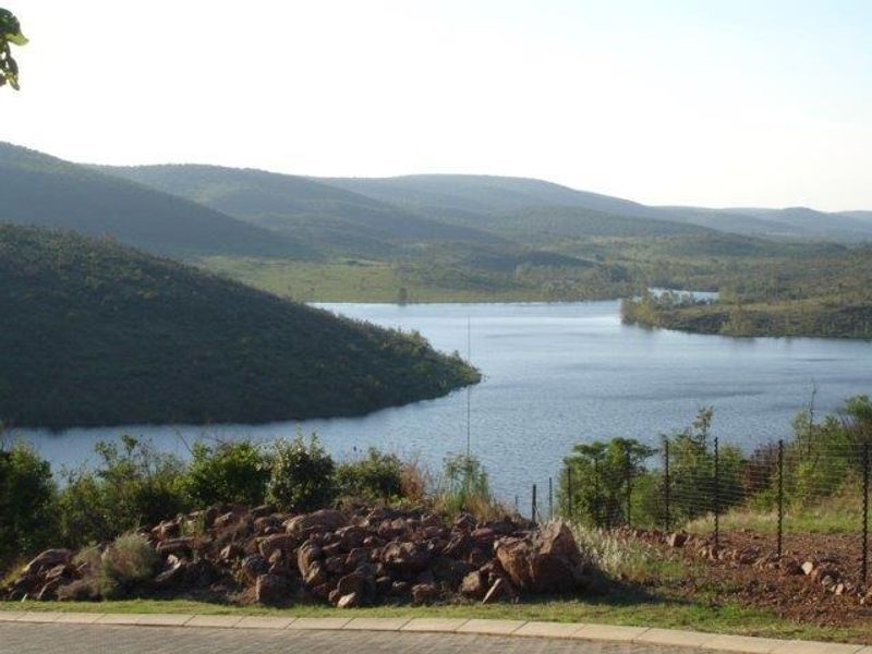 A scenic view overlooking a large dam or lake surrounded by rolling hills and natural vegetation, captured from the edge of a residential property.