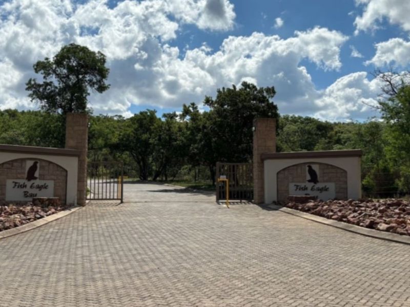 The entrance to Fish Eagle Bay featuring a paved driveway, secure gated access, and stone masonry pillars set within a natural landscape.