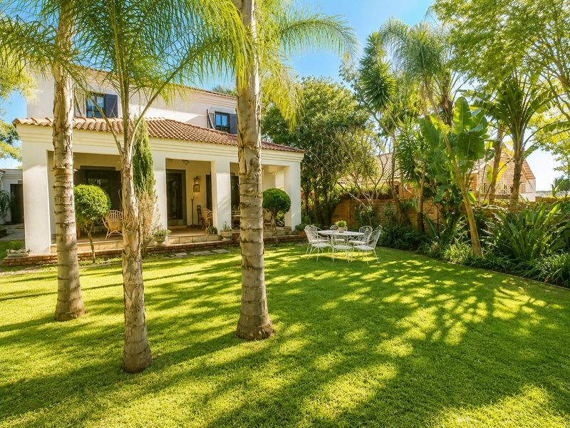 A well-maintained rear garden featuring a lush lawn, mature palm trees, and a covered patio area attached to the main house structure.