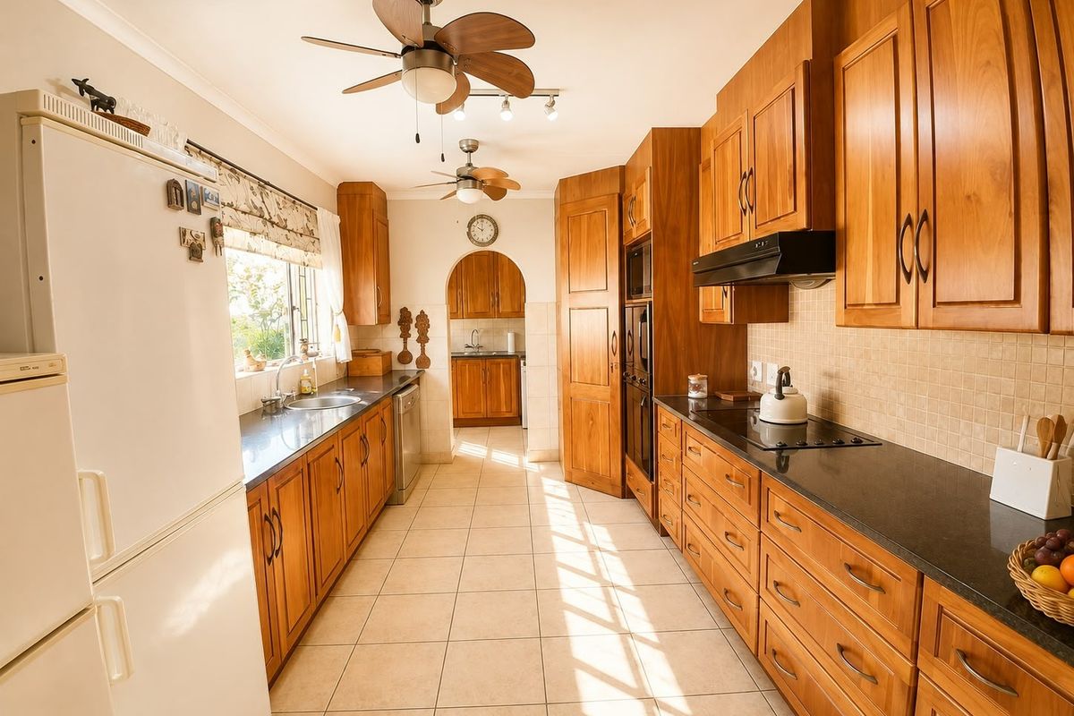 A spacious kitchen featuring extensive wooden cabinetry, granite countertops, built-in appliances, and tiled flooring, with an adjoining scullery area visible through an arched doorway.