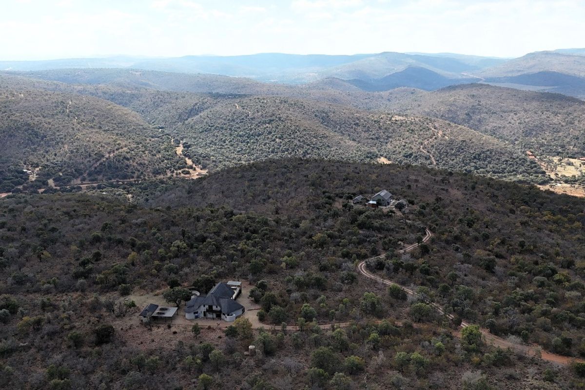 An aerial view of a residential property situated within a vast, natural bushveld landscape, featuring an unpaved access road and surrounding hilly terrain.