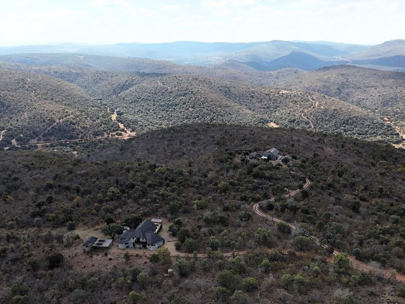 An aerial view of a residential property situated within a vast, natural bushveld landscape, featuring an unpaved access road and surrounding hilly terrain.