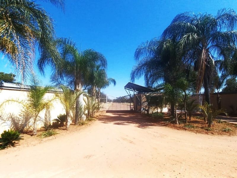 A view of the property entrance featuring a security gate, guard house, perimeter wall, and an unpaved driveway lined with palm trees.