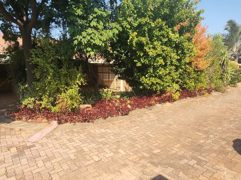 A view of the townhouse front exterior featuring a paved driveway, established garden beds, and mature trees providing shade to the brick facade.