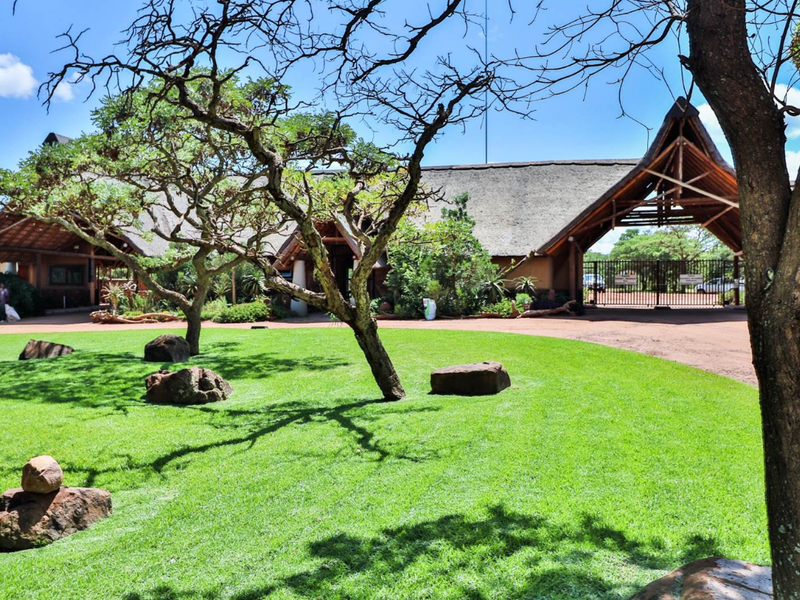 A well-maintained front entrance featuring a traditional thatched roof structure, gated access, and a manicured lawn with mature trees.
