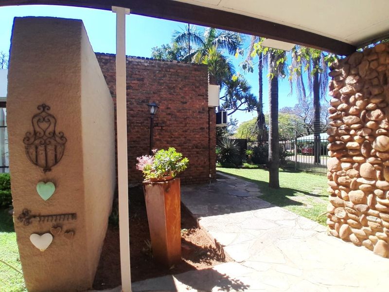 A covered patio area featuring stone and brick wall finishes, overlooking a landscaped garden with paved pathways and secure perimeter fencing.