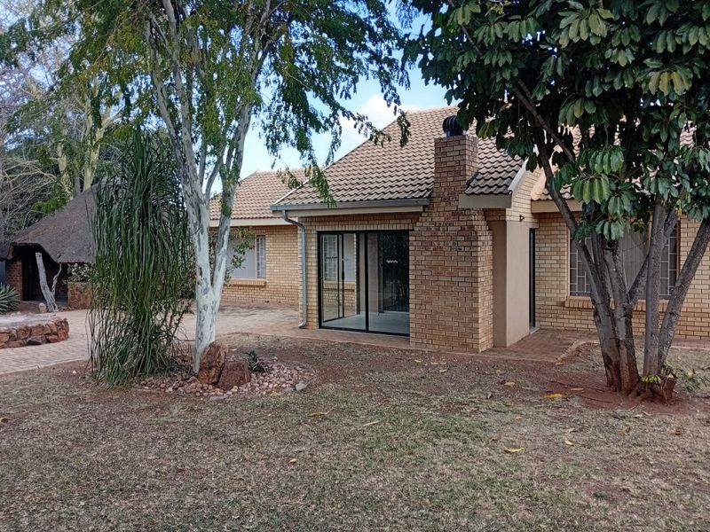 Rear view of a brick house featuring a tiled roof, brick chimney, sliding glass doors leading to a paved patio, and a separate thatched lapa.