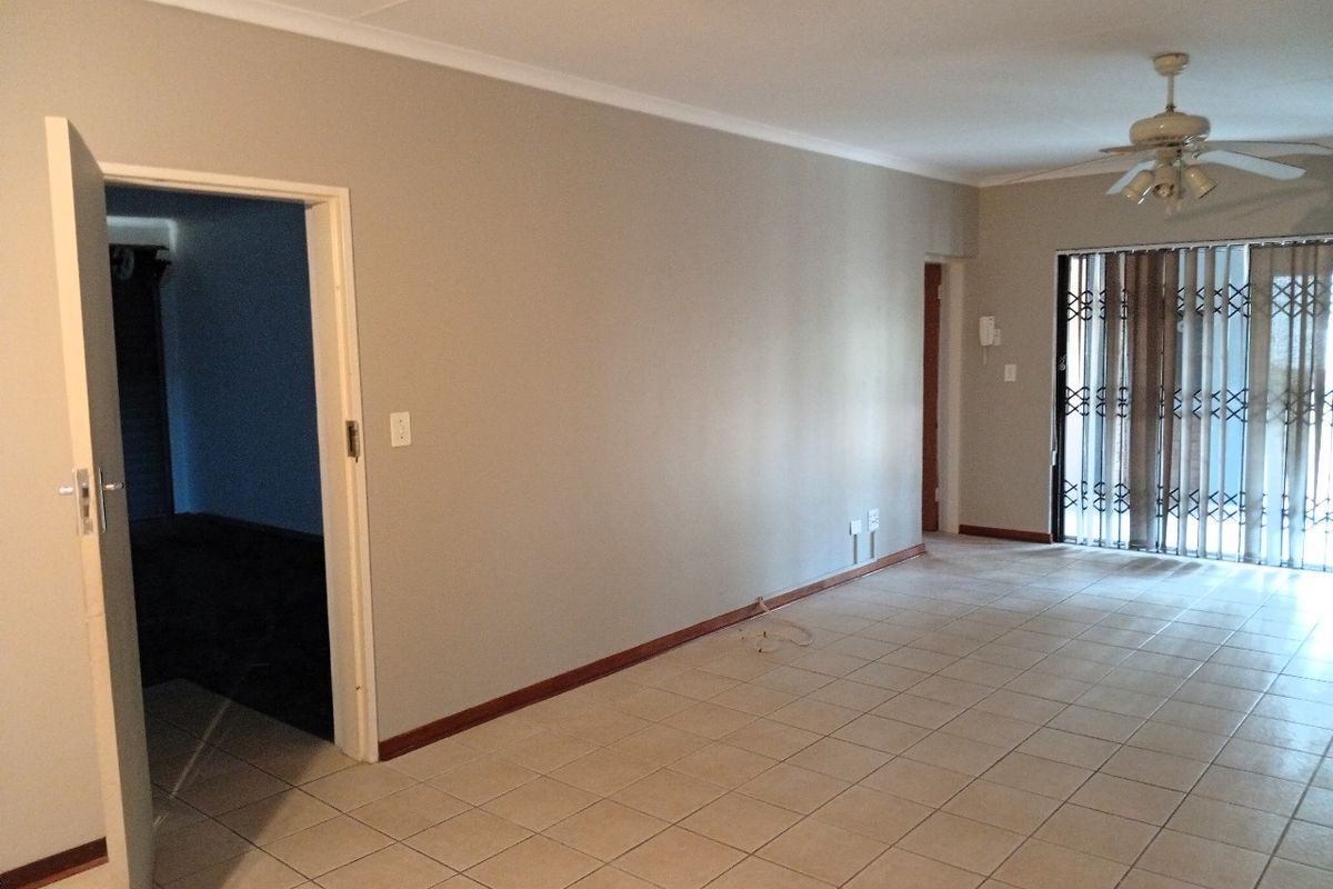 A spacious, empty living area featuring neutral walls, tiled flooring, a ceiling fan, and a sliding door with a security gate.