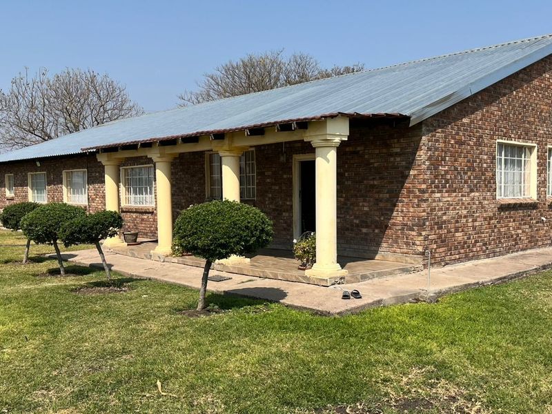 A single-story face brick home featuring a covered patio with concrete columns, set within a spacious garden area.