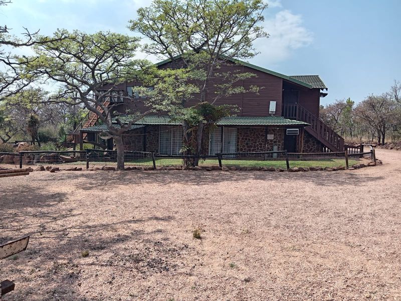 A two-story house featuring stone and timber cladding, a tiled roof, and an external staircase, set within a natural bushveld environment.