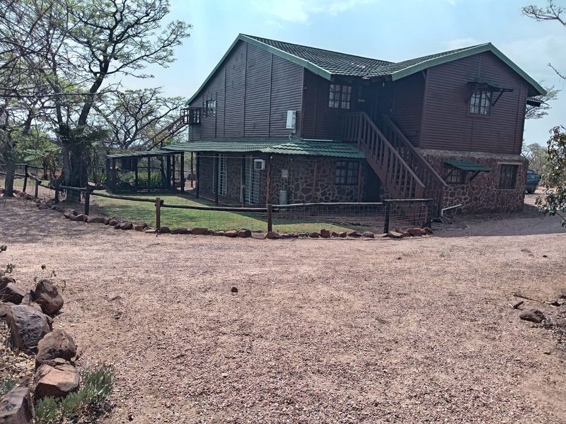 A two-story house featuring timber cladding and stone masonry, with an external staircase and surrounding gravel driveway in a natural setting.