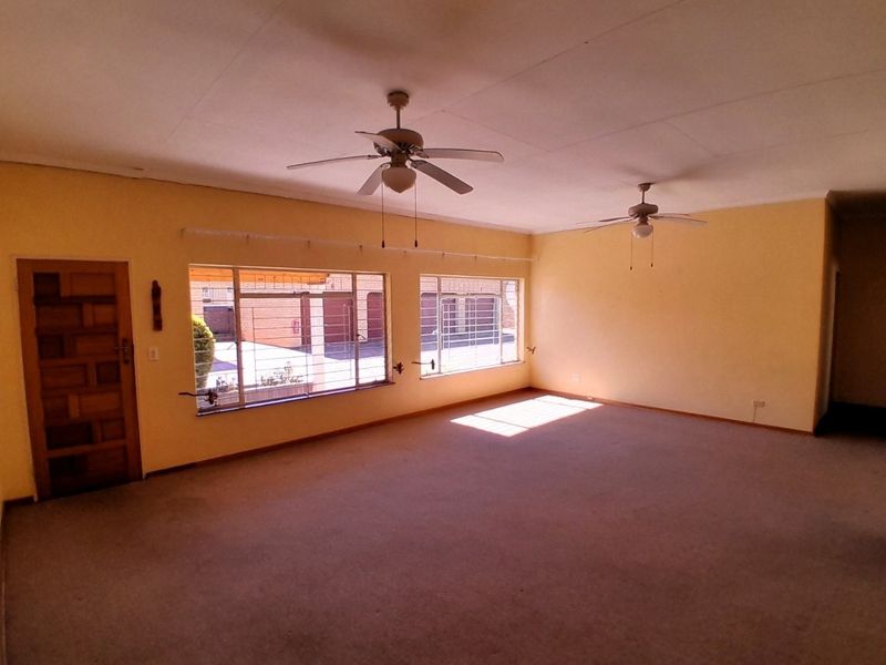 Spacious, empty living room featuring dual ceiling fans, large windows with security bars, and carpeted flooring, offering a blank canvas for renovation.