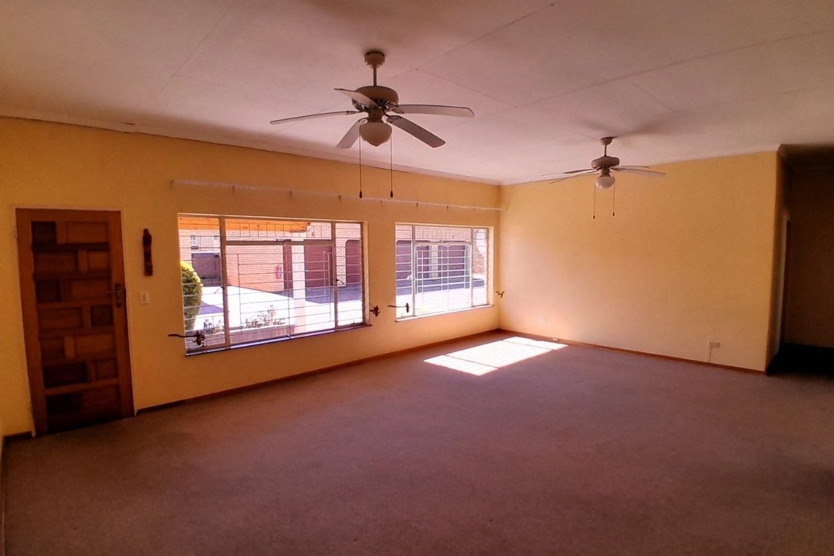 Spacious, empty living room featuring dual ceiling fans, large windows with security bars, and carpeted flooring, offering a blank canvas for renovation.