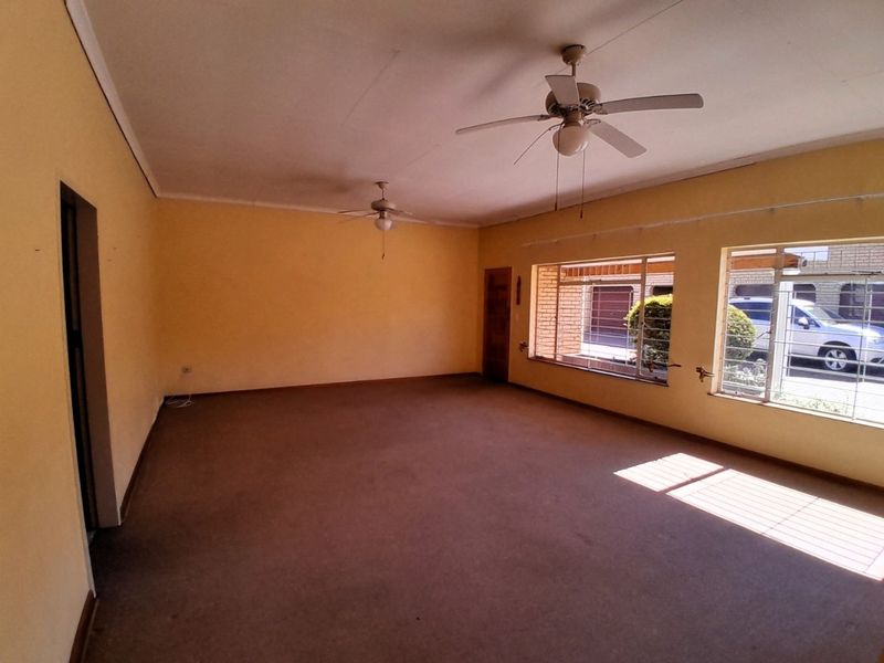 Spacious, empty living area featuring dual ceiling fans, carpeted flooring, and large windows with security bars, offering a blank canvas for renovation.