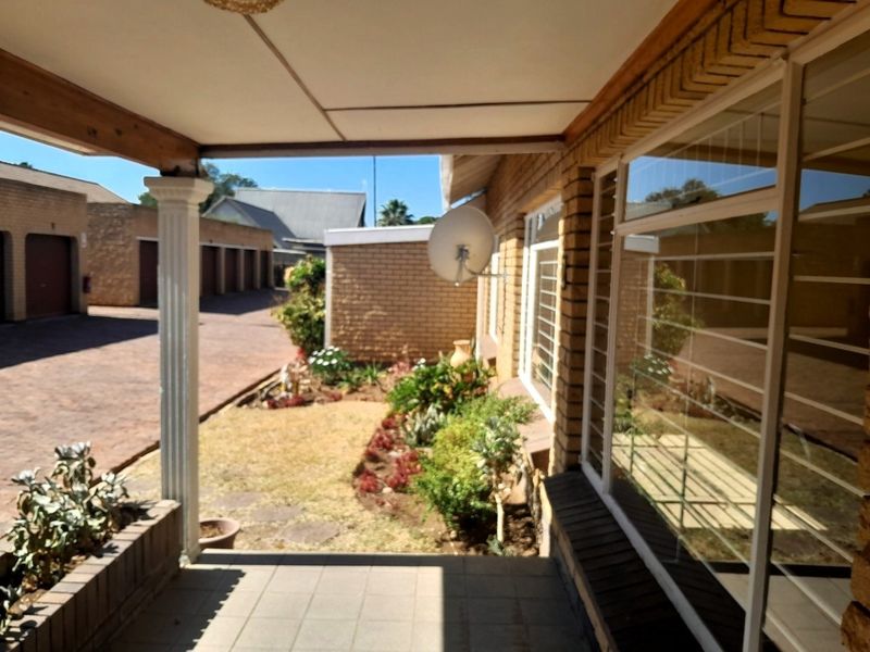 A covered patio area featuring tiled flooring and a decorative column, overlooking a small garden and the townhouse complex driveway.