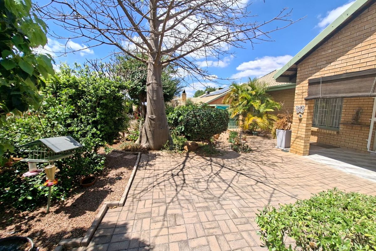 A well-maintained rear garden area featuring brick paving, established trees, and a face brick exterior facade with security bars on the windows.