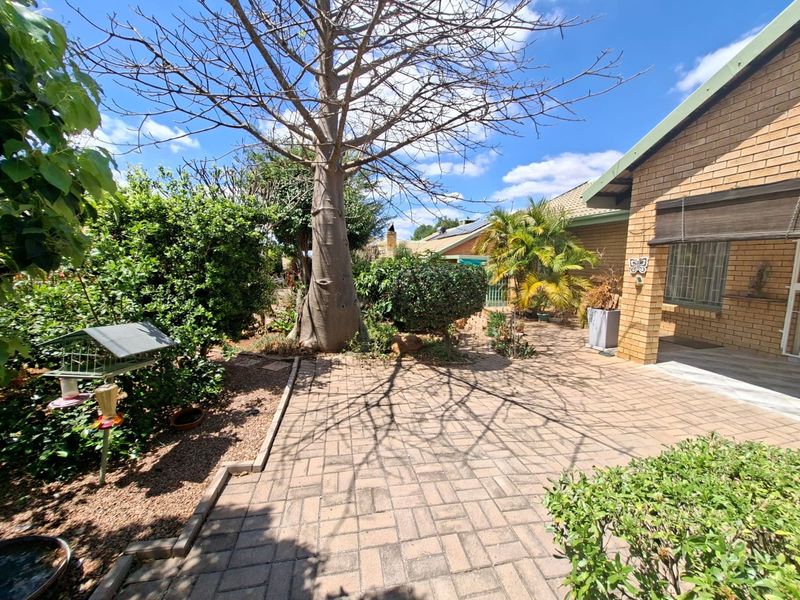 A well-maintained rear garden area featuring brick paving, established trees, and a face brick exterior facade with security bars on the windows.