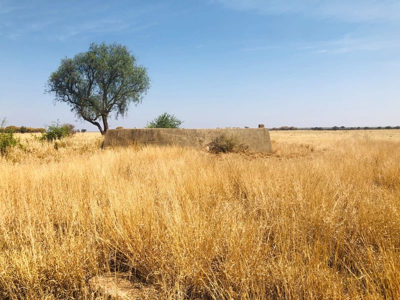 A view of a vacant residential plot featuring dry grass and an old, weathered concrete water reservoir structure.