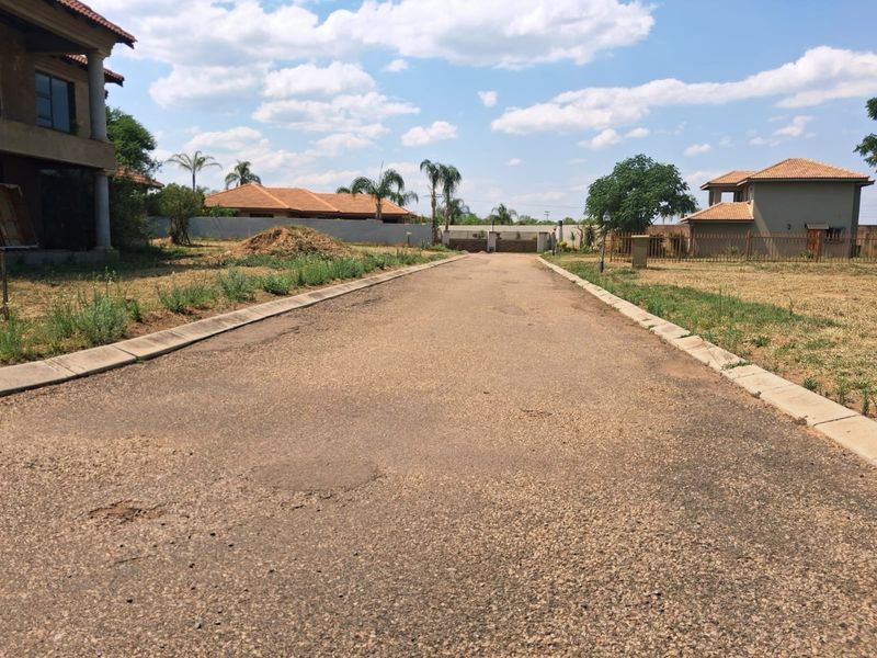 A view of a paved residential street with concrete curbing, showing surrounding properties and perimeter fencing in a suburban estate setting.