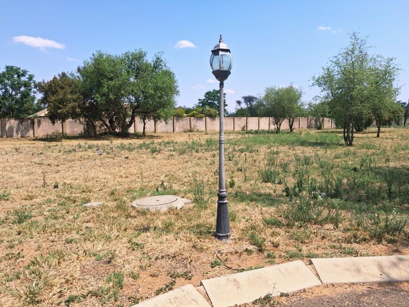 A vacant residential plot featuring a perimeter wall, a street lamp, and a manhole cover, set within a grassy landscape.