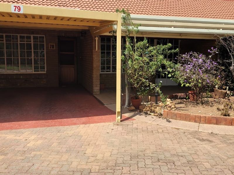 Front exterior view of a townhouse featuring a covered carport, brick facade, tiled roof, and a paved driveway with a small garden area.