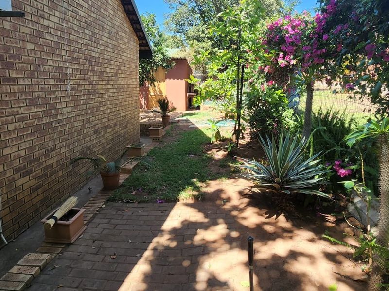 A side garden area featuring brick paving, a facebrick exterior wall, and established garden beds with mature plants and fencing.