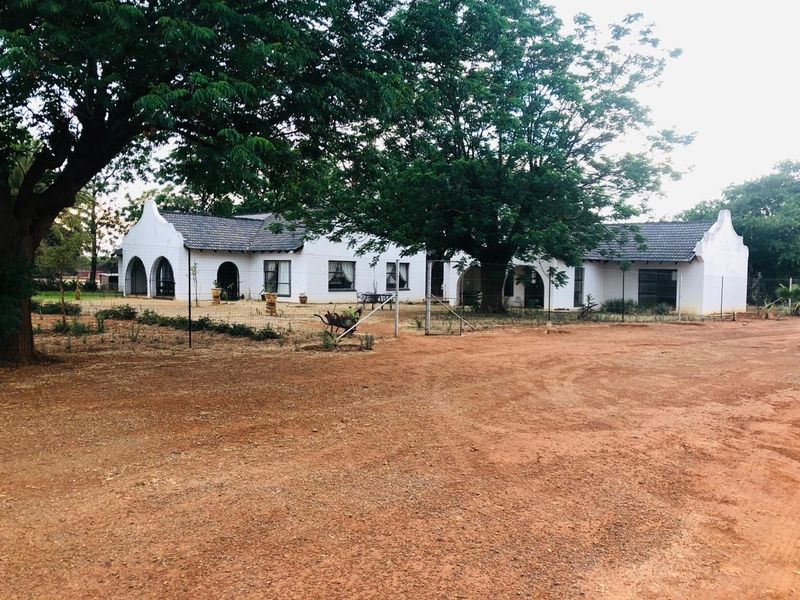 A front view of a single-story small holding residence featuring a gabled roof, arched patio areas, and a large surrounding garden with mature trees.