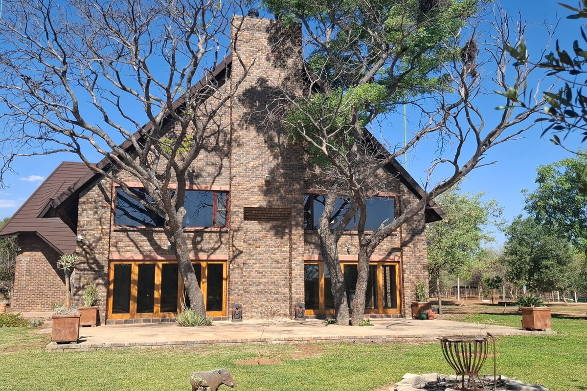 A rear view of a double-story brick farmhouse featuring a prominent central chimney, large glass doors opening onto a paved patio, and mature trees.