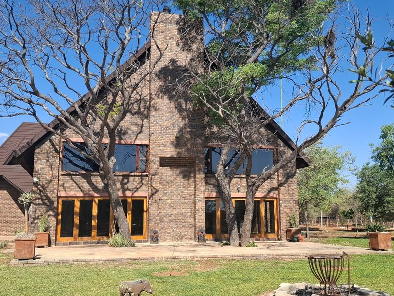 A rear view of a double-story brick farmhouse featuring a prominent central chimney, large glass doors opening onto a paved patio, and mature trees.