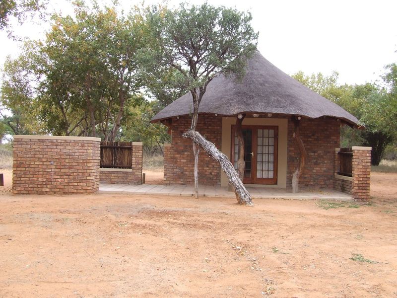 A charming face brick cottage with a traditional thatched roof, featuring a paved patio area and french door entrance in a natural bushveld setting.