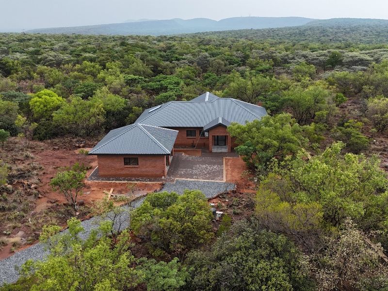 Aerial view of a modern brick house with a metal roof, situated in a natural bushveld setting with a paved driveway and patio area.