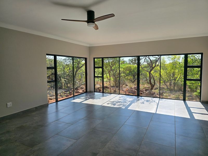 A bright, empty living room featuring large aluminium-framed windows with natural bushveld views, modern tiled flooring, and a ceiling fan.