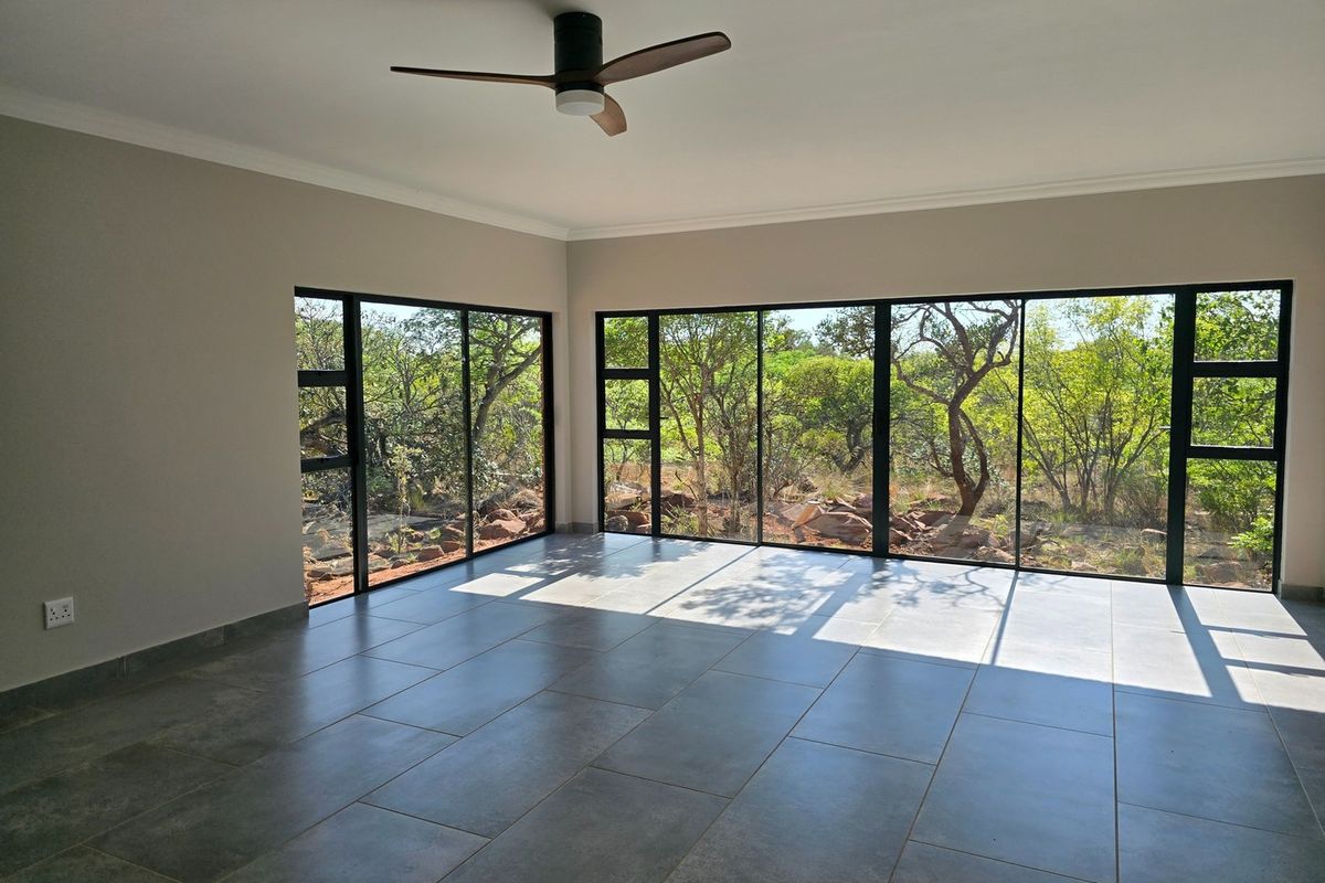 A bright, empty living room featuring large aluminium-framed windows with natural bushveld views, modern tiled flooring, and a ceiling fan.