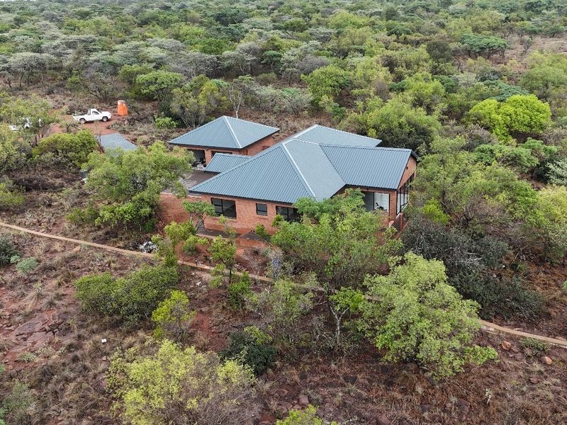 Aerial view of a brick house with a metal roof, situated in a natural, wooded landscape.