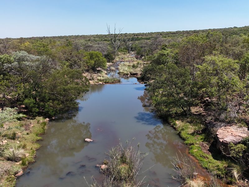 A scenic aerial view of a natural river flowing through the farm landscape, surrounded by indigenous trees and rocky terrain.