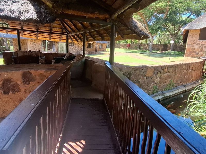 A rustic outdoor patio area featuring a thatched roof, stone masonry walls, and a wooden footbridge overlooking a tranquil pond.