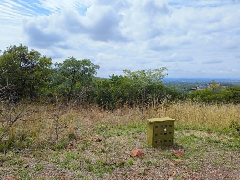 A view of a vacant residential plot featuring natural vegetation, dry grass, and a utility box, set against a scenic landscape background.