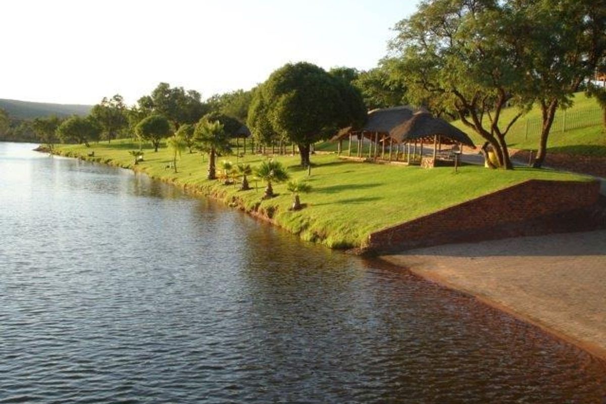 Scenic waterfront view featuring a well-maintained lawn, mature trees, a brick retaining wall, and a thatched lapa structure along the shoreline.