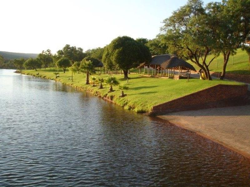 Scenic waterfront view featuring a well-maintained lawn, mature trees, a brick retaining wall, and a thatched lapa structure along the shoreline.