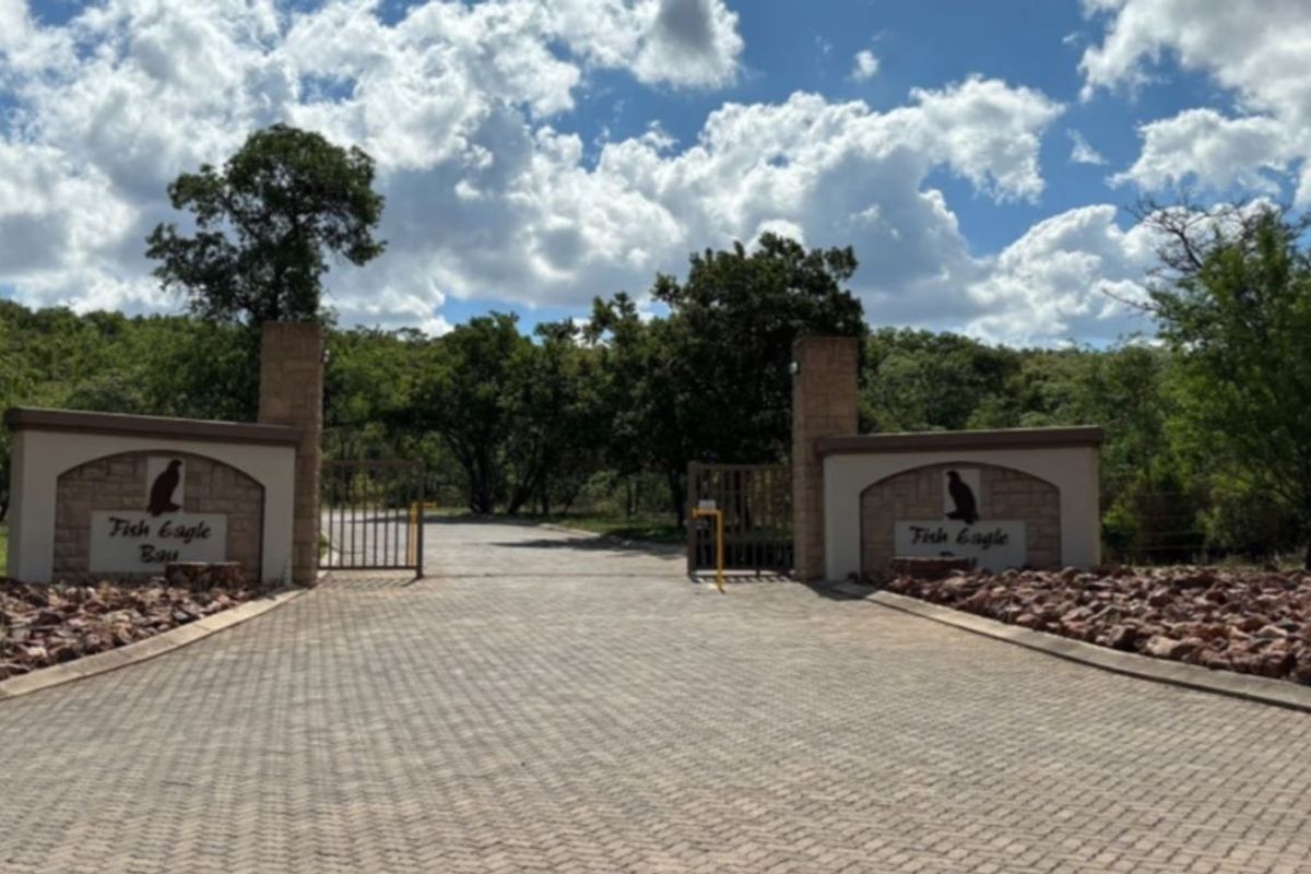 The entrance to Fish Eagle Bay featuring a paved driveway, secure gated access, and stone masonry pillars set within a natural landscape.