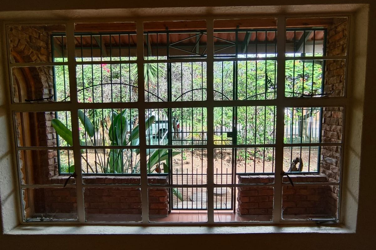 Interior view looking through a large window with security bars, featuring exposed brick walls and a view of the garden area.