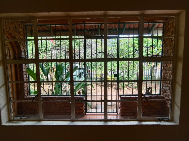 Interior view looking through a large window with security bars, featuring exposed brick walls and a view of the garden area.