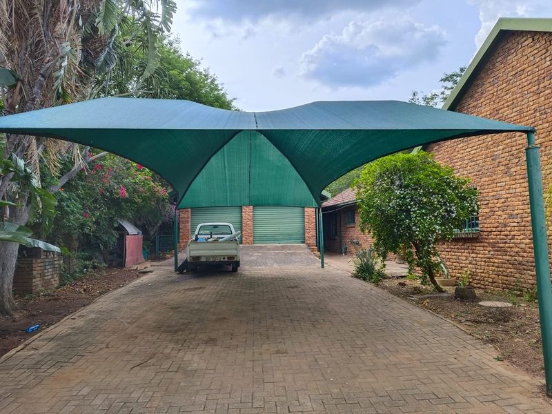 Exterior view of a driveway featuring a large green shade cloth carport, face brick facade, and double roller shutter garage doors.