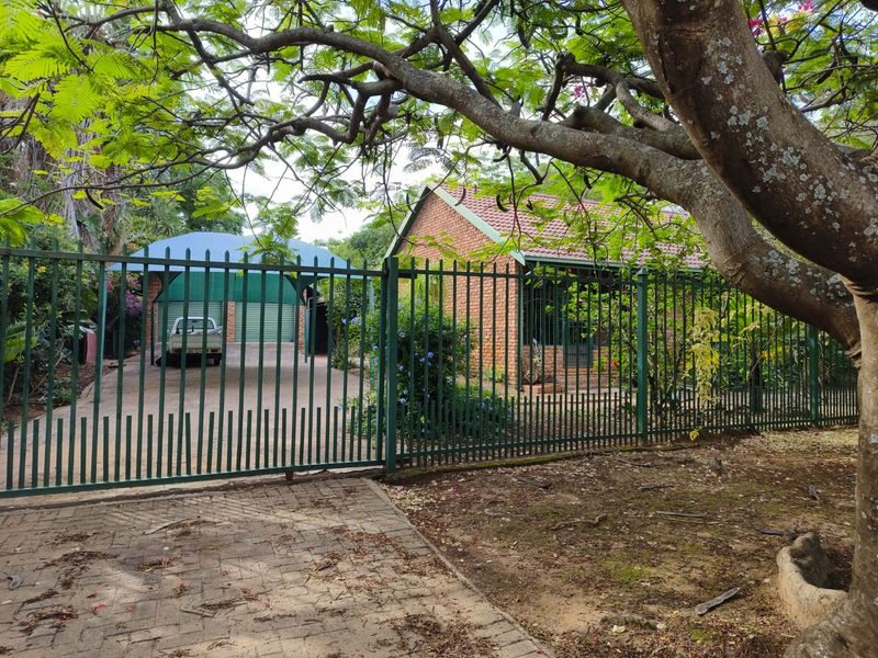 Front view of a brick house featuring a paved driveway, carport, and secure metal perimeter fencing surrounded by mature trees.