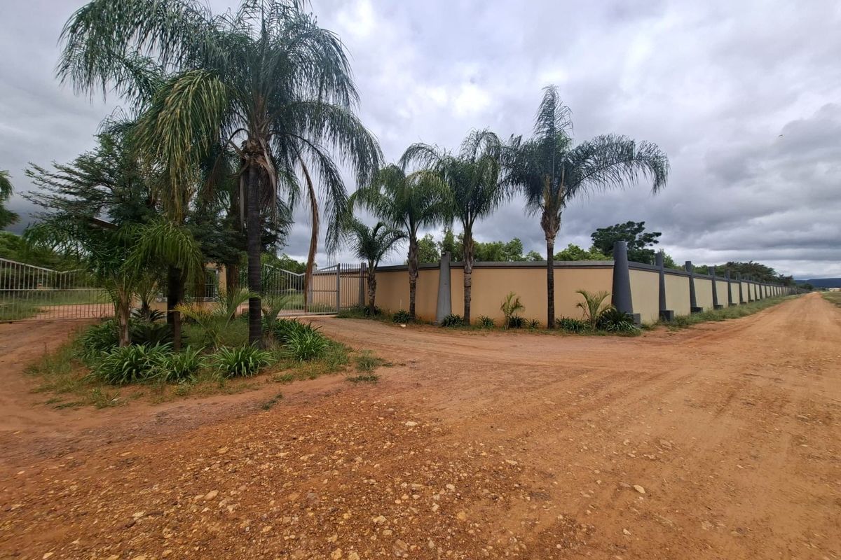 A view of the property entrance featuring a secure perimeter wall, metal gate, and established palm trees along a dirt access road.
