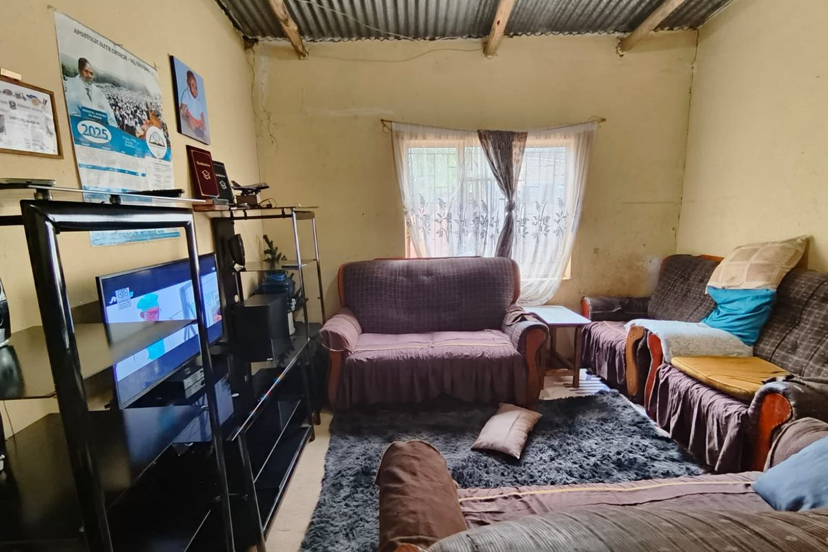 A living room featuring exposed timber rafters and a corrugated metal ceiling, with visible wall surface wear and security bars on the window.
