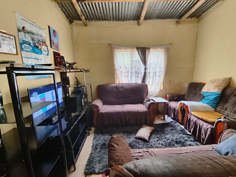A living room featuring exposed timber rafters and a corrugated metal ceiling, with visible wall surface wear and security bars on the window.